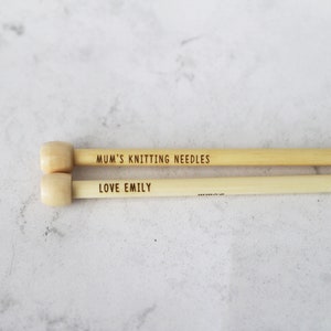 May include: Two light-colored wooden knitting needles with rounded ends. The top needle reads "MUM'S KNITTING NEEDLES" and the bottom needle reads "LOVE EMILY". The needles are lying on a light gray surface.
