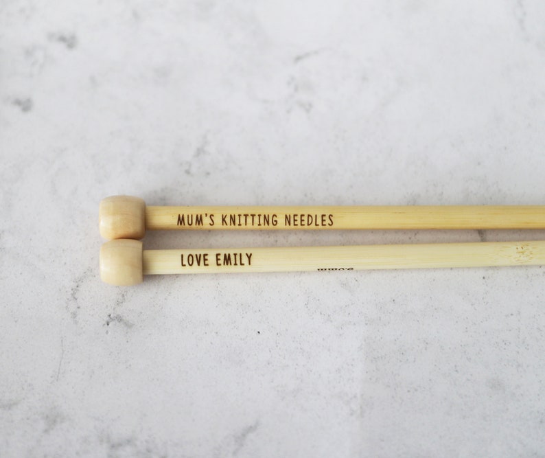 May include: Two light-colored wooden knitting needles with rounded ends. The top needle reads "MUM'S KNITTING NEEDLES" and the bottom needle reads "LOVE EMILY". The needles are lying on a light gray surface.