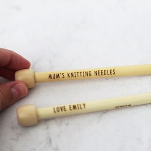 May include: Two wooden knitting needles with light-colored wooden tips. One needle is engraved with "MUM'S KNITTING NEEDLES," and the other with "LOVE EMILY." The needles are light beige in color and are likely used for crafting.