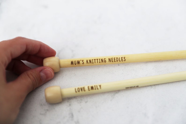 May include: Two wooden knitting needles with light-colored wooden tips. One needle is engraved with "MUM'S KNITTING NEEDLES," and the other with "LOVE EMILY." The needles are light beige in color and are likely used for crafting.
