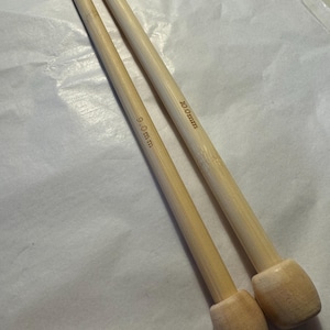 May include: Two bamboo knitting needles. One needle is marked "9.0mm" and the other is marked "10.0mm". The needles are light brown with rounded ends. The background is a light-colored, textured surface.