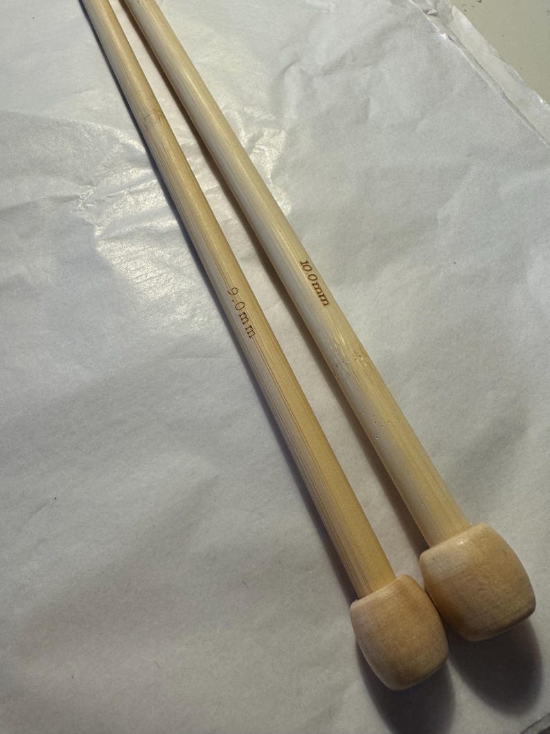May include: Two bamboo knitting needles. One needle is marked "9.0mm" and the other is marked "10.0mm". The needles are light brown with rounded ends. The background is a light-colored, textured surface.
