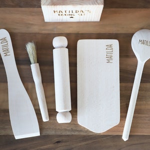 May include: A wooden baking set with the name "MATILDA" engraved on a spatula, spoon, and cutting board. The set includes a rolling pin, brush, and a small wooden box with "MATILDA'S BAKING SET" engraved on it. The items are arranged on a wooden surface.