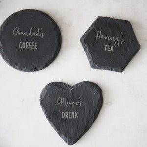 Puede incluir: Tres posavasos de pizarra negra con texto grabado. El primer posavasos es redondo y dice "Grandad's Coffee". El segundo posavasos es un hex&aacute;gono y dice "Nanny's Tea". El tercer posavasos tiene forma de coraz&oacute;n y dice "Mumi's Drink".