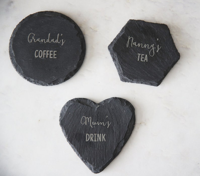 Puede incluir: Tres posavasos de pizarra negra con texto grabado. El primer posavasos es redondo y dice "Grandad's Coffee". El segundo posavasos es un hex&aacute;gono y dice "Nanny's Tea". El tercer posavasos tiene forma de coraz&oacute;n y dice "Mumi's Drink".