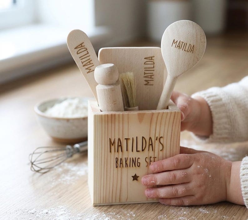 May include: A wooden baking set for children, featuring a box with the text "MATILDA'S BAKING SET" and a star. The set includes a spoon, spatula, brush, and rolling pin, each engraved with the name "MATILDA".
