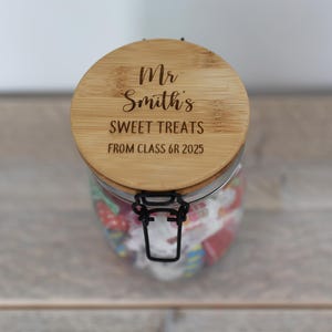 May include: A glass jar filled with sweets, featuring a wooden lid engraved with "Mr Smith's SWEET TREATS FROM CLASS 6R 2025". The jar has a metal clasp. The jar is filled with colorful sweets.