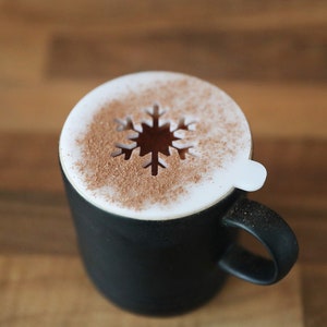 May include: A black mug with a white snowflake design on top. The snowflake is made of cinnamon.