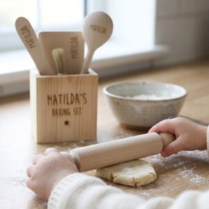 May include: A wooden baking set with the text "MATILDA'S BAKING SET" and a star. The set includes a wooden spoon, spatula, and brush, all with the name "MATILDA" engraved. A child is rolling dough with a wooden rolling pin. A bowl of flour is nearby.