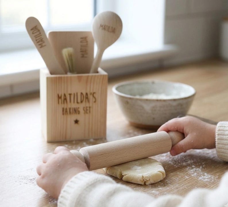 May include: A wooden baking set with the text "MATILDA'S BAKING SET" and a star. The set includes a wooden spoon, spatula, and brush, all with the name "MATILDA" engraved. A child is rolling dough with a wooden rolling pin. A bowl of flour is nearby.