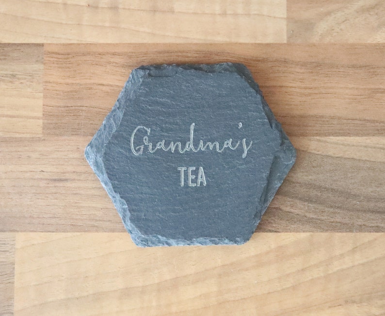 Puede incluir: Un posavasos hexagonal de pizarra con el texto "Grandma's Tea" grabado en blanco.