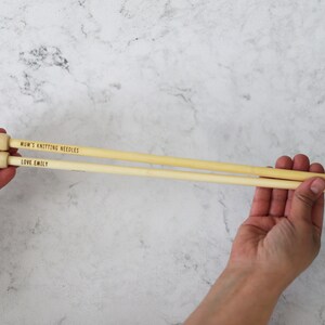 May include: Two light-colored wooden knitting needles are held in a person's hands. The needles have rounded tops and pointed ends. Text on the needles reads "MUM'S KNITTING NEEDLES" and "LOVE EMILY".