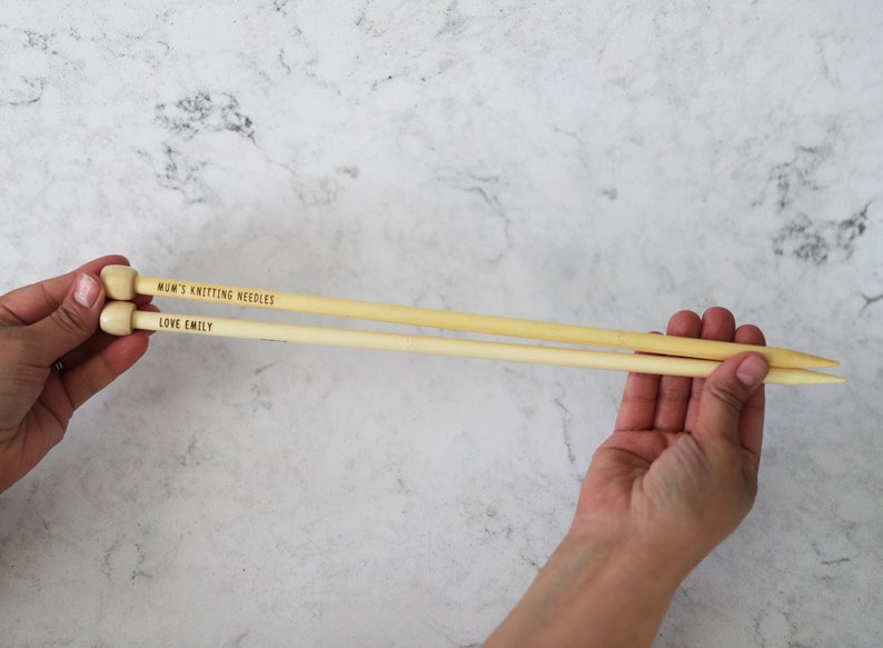 May include: Two light-colored wooden knitting needles are held in a person's hands. The needles have rounded tops and pointed ends. Text on the needles reads "MUM'S KNITTING NEEDLES" and "LOVE EMILY".