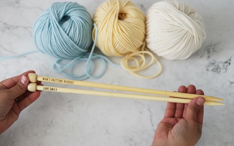 May include: A pair of wooden knitting needles with the text "Mum's Knitting Needles" and "Love Emily" are held in a person's hands. Three balls of yarn in light blue, yellow, and white are in the background. The needles are a light beige color.