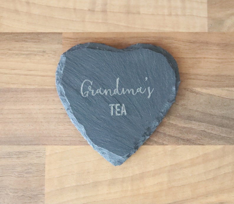 Puede incluir: Un posavasos de pizarra en forma de coraz&oacute;n con el texto "Grandma's Tea" grabado en &eacute;l.