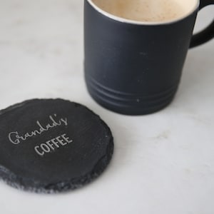 Puede incluir: Un posavasos de pizarra negro con el texto "Granddad's Coffee" grabado en &eacute;l. El posavasos est&aacute; sobre una superficie blanca junto a una taza de caf&eacute; negra.
