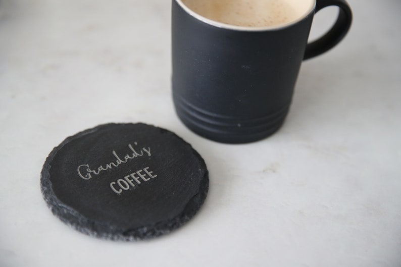 Puede incluir: Un posavasos de pizarra negro con el texto "Granddad's Coffee" grabado en &eacute;l. El posavasos est&aacute; sobre una superficie blanca junto a una taza de caf&eacute; negra.