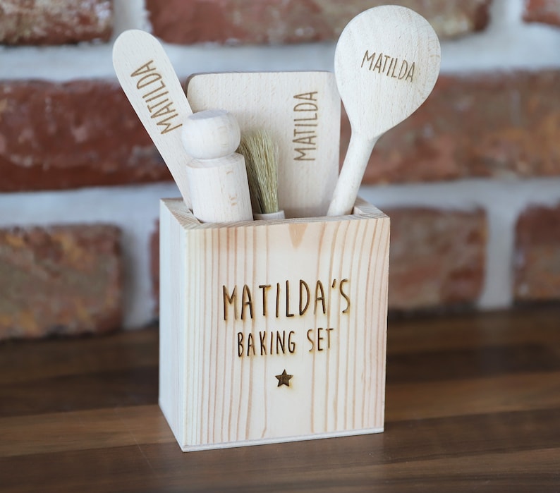 May include: A wooden baking set for Matilda, featuring a square container with "Matilda's Baking Set" engraved on the front. The set includes a spatula, a rolling pin, a brush, and a spoon, each engraved with "Matilda". The set is made of light-colored wood.