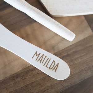 May include: Wooden kitchen utensils, including a spatula engraved with the name "MATILDA", a rolling pin, and a cutting board. The utensils are light-colored and set against a dark wood background.
