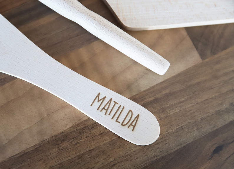 May include: Wooden kitchen utensils, including a spatula engraved with the name "MATILDA", a rolling pin, and a cutting board. The utensils are light-colored and set against a dark wood background.