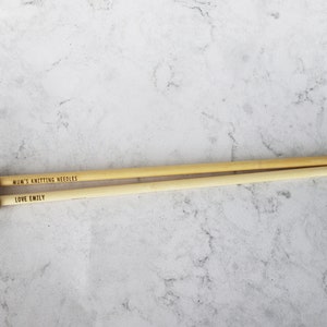 May include: Two wooden knitting needles with rounded ends and pointed tips. The text "MUM'S KNITTING NEEDLES" and "LOVE EMILY" are engraved on the needles. The needles are light brown and lie on a light gray and white marble surface.