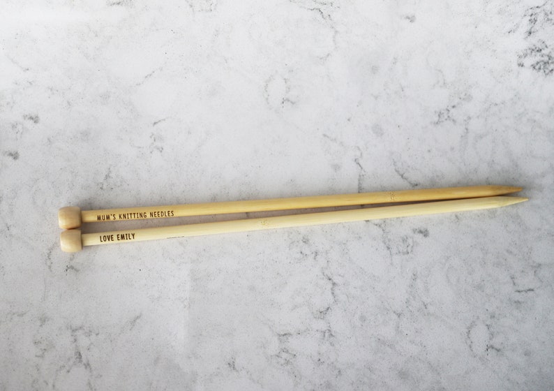 May include: Two wooden knitting needles with rounded ends and pointed tips. The text "MUM'S KNITTING NEEDLES" and "LOVE EMILY" are engraved on the needles. The needles are light brown and lie on a light gray and white marble surface.