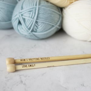 May include: Close-up of knitting needles and yarn balls. The wooden needles are engraved with "MUM'S KNITTING NEEDLES" and "LOVE EMILY". Several balls of yarn in shades of blue and cream are in the background. The image is well-lit and shows a craft theme.