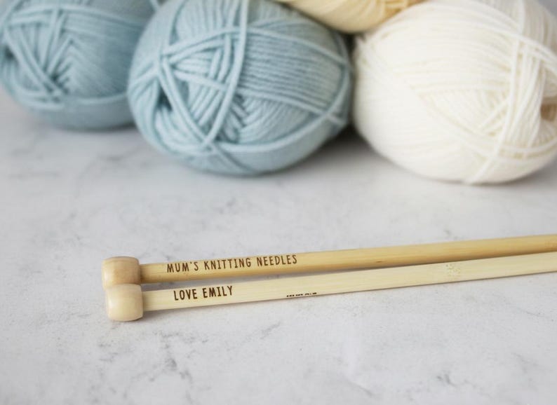 May include: Close-up of knitting needles and yarn balls. The wooden needles are engraved with "MUM'S KNITTING NEEDLES" and "LOVE EMILY". Several balls of yarn in shades of blue and cream are in the background. The image is well-lit and shows a craft theme.