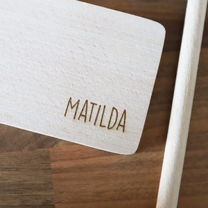 May include: A light-colored wooden cutting board with the name "MATILDA" engraved on it. A wooden handle is attached to the board. The board is made of light-colored wood.