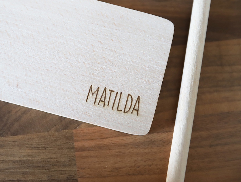 May include: A light-colored wooden cutting board with the name "MATILDA" engraved on it. A wooden handle is attached to the board. The board is made of light-colored wood.
