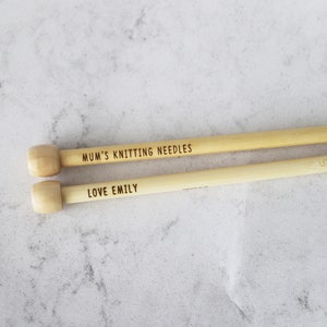 May include: Two wooden knitting needles with light-colored wooden knobs. The top needle reads "MUM'S KNITTING NEEDLES" and the bottom needle reads "LOVE EMILY". The needles are a light tan color.
