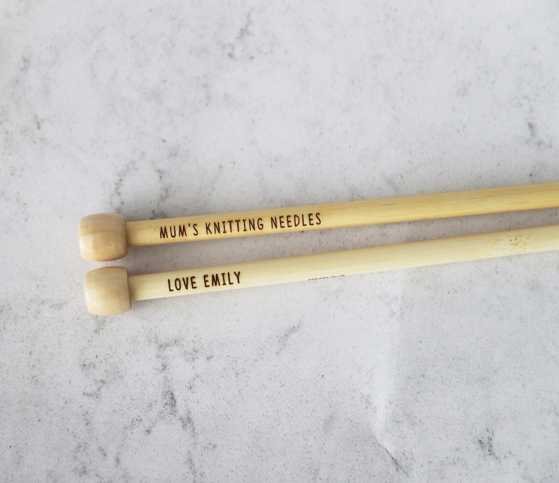 May include: Two wooden knitting needles with light-colored wooden knobs. The top needle reads "MUM'S KNITTING NEEDLES" and the bottom needle reads "LOVE EMILY". The needles are a light tan color.