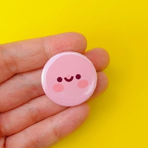May include: A pink button badge with a smiling face design. The badge has a light pink background with a simple cartoon face featuring black eyes and a curved mouth. The badge is held against a bright yellow background.