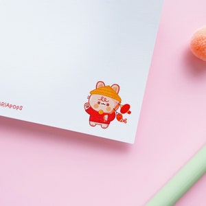 May include: A cute cartoon illustration of a cat wearing a red and yellow outfit and a yellow hat. The cat is holding a red lantern and a red fish. The illustration is on a white background.
