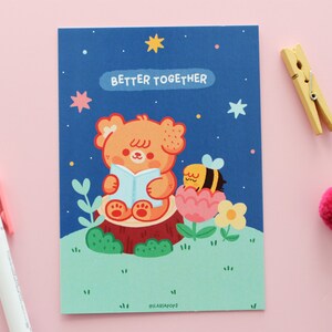 May include: A blue card with a cartoon illustration of a bear reading a book and a bee sitting in a flower. The text "Better Together" is written in white on the card.
