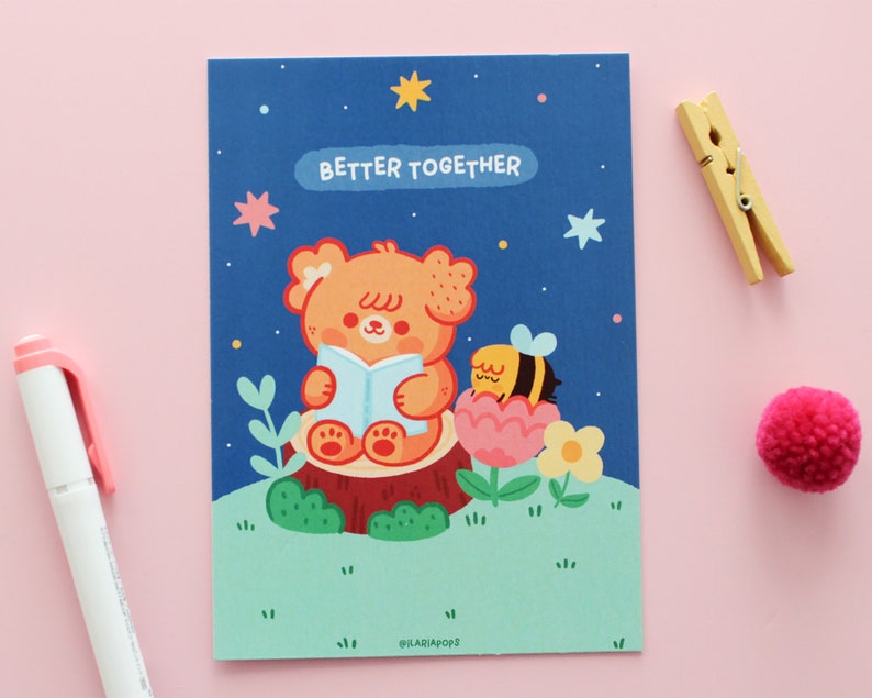 May include: A blue card with a cartoon illustration of a bear reading a book and a bee sitting in a flower. The text "Better Together" is written in white on the card.