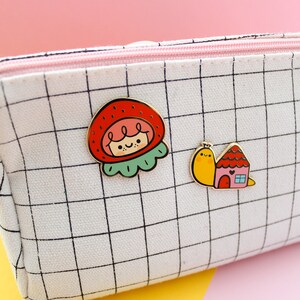 May include: White canvas pencil case with a black grid pattern. Two enamel pins are attached to the case. One pin is a red and green strawberry with a smiling face. The other pin is a yellow snail carrying a pink and red house.