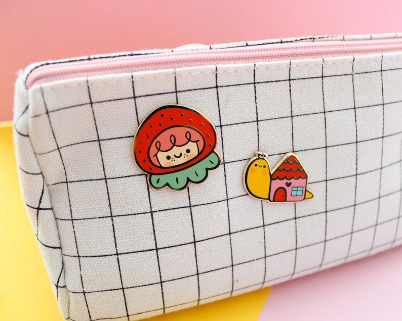 May include: White canvas pencil case with a black grid pattern. Two enamel pins are attached to the case. One pin is a red and green strawberry with a smiling face. The other pin is a yellow snail carrying a pink and red house.
