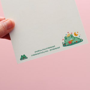 May include: A white card with a green frog illustration on the bottom left corner. The frog is wearing a red bow tie and has the text "ILARIAPOPS!" printed on it. The top right corner of the card features a green frog with a yellow star on its head, sleeping on a bed of flowers. The frog is holding a cup of tea. The text "PATREON.COM/ILARIAPOPS ILARIAPOPS.ETSY.COM - @ILARIAPOPS" is printed on the bottom right corner of the card.