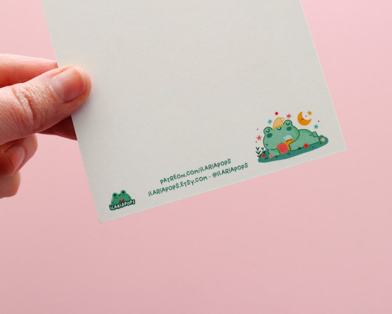 May include: A white card with a green frog illustration on the bottom left corner. The frog is wearing a red bow tie and has the text "ILARIAPOPS!" printed on it. The top right corner of the card features a green frog with a yellow star on its head, sleeping on a bed of flowers. The frog is holding a cup of tea. The text "PATREON.COM/ILARIAPOPS ILARIAPOPS.ETSY.COM - @ILARIAPOPS" is printed on the bottom right corner of the card.