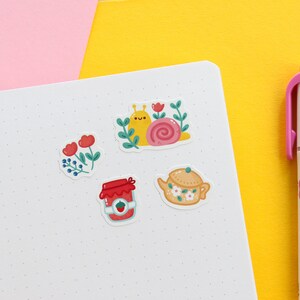 Pode incluir: Um conjunto de cinco adesivos com ilustra&ccedil;&otilde;es fofas de um caracol, um bule, um pote de geleia e dois arranjos florais. Os adesivos est&atilde;o em um fundo branco com um padr&atilde;o de pontos sutil.