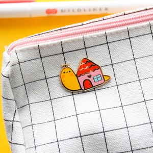 May include: A yellow and red enamel pin featuring a snail carrying a small house with a pink door and a window. The pin is on a white canvas zippered pouch with a black grid pattern.