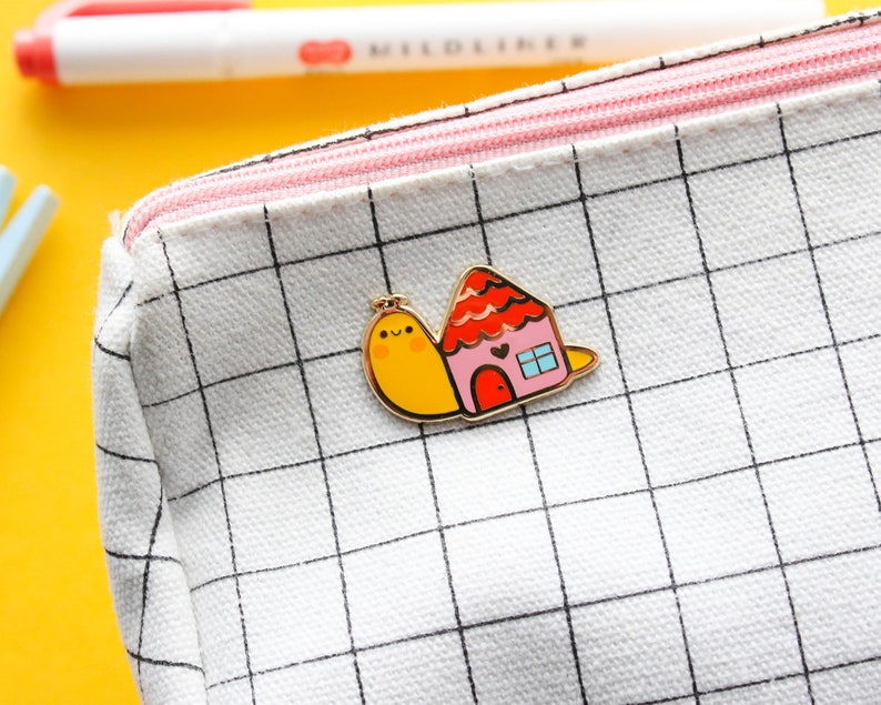 May include: A yellow and red enamel pin featuring a snail carrying a small house with a pink door and a window. The pin is on a white canvas zippered pouch with a black grid pattern.