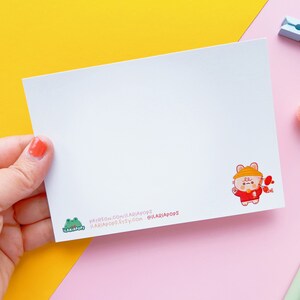 May include: A white card featuring a cartoon illustration of a brown and white animal wearing a red and white hat and holding a red and white fish. The card has text at the bottom that reads "patreon.com/ilariadops ilariadops.etsy.com @ilariadops".
