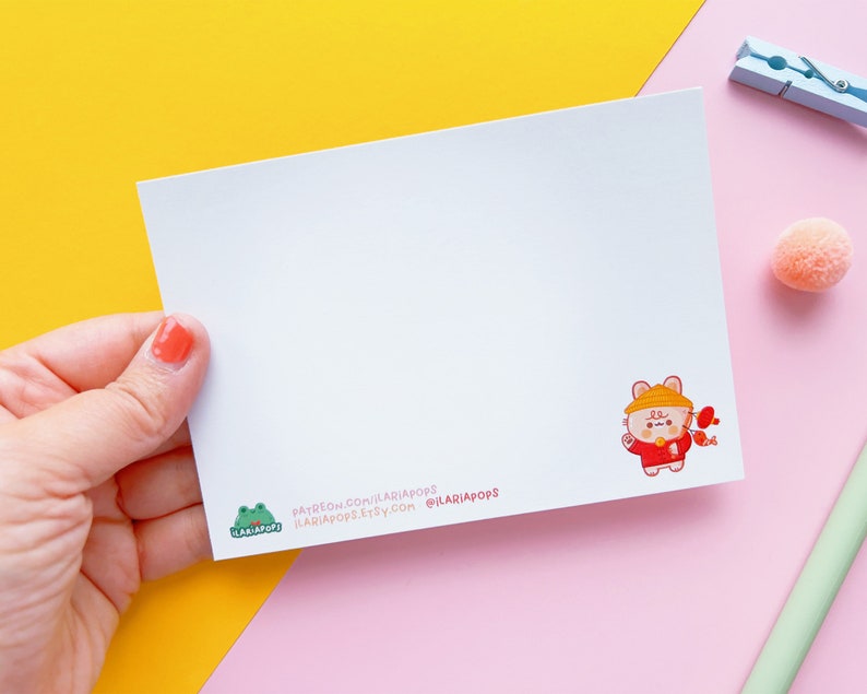 May include: A white card featuring a cartoon illustration of a brown and white animal wearing a red and white hat and holding a red and white fish. The card has text at the bottom that reads "patreon.com/ilariadops ilariadops.etsy.com @ilariadops".