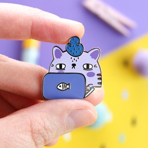 Può includere: Spilla smaltata a forma di gatto viola con un fiore blu sulla testa, che indossa un laptop blu con un'icona di pesce bianco sullo schermo. Il gatto sta guardando il laptop.