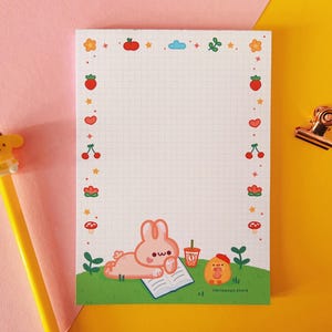 Puede incluir: Bloc de notas con un patrón de cuadrícula y un borde de frutas y flores ilustradas. El bloc de notas presenta un conejo de dibujos animados, una bebida y un pollito. También se ve un bolígrafo amarillo con un personaje de dibujos animados y un clip dorado.
