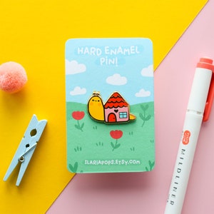 May include: A yellow snail carrying a small pink house with a red door. The enamel pin is on a white card with blue clouds and green grass. The card reads "Hard Enamel Pin!" and "ilariapops.etsy.com".