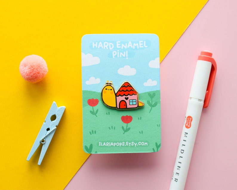 May include: A yellow snail carrying a small pink house with a red door. The enamel pin is on a white card with blue clouds and green grass. The card reads "Hard Enamel Pin!" and "ilariapops.etsy.com".