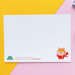 May include: A white card featuring a charming cartoon illustration of a bear dressed in a red and yellow outfit, holding a red fish. The card includes the text "PATREON.COM/ILARIAPOPS ILARIAPOPS.ETSY.COM @ILARIAPOPS" printed at the bottom.
